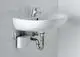 Weißes Wandwaschbecken mit einem grauen CLAGE E-Kleindurchlauferhitzer darunter, verbunden durch eine metallene Abflussleitung und Wasseranschlüsse, an einer hellen Fliesenwand montiert.